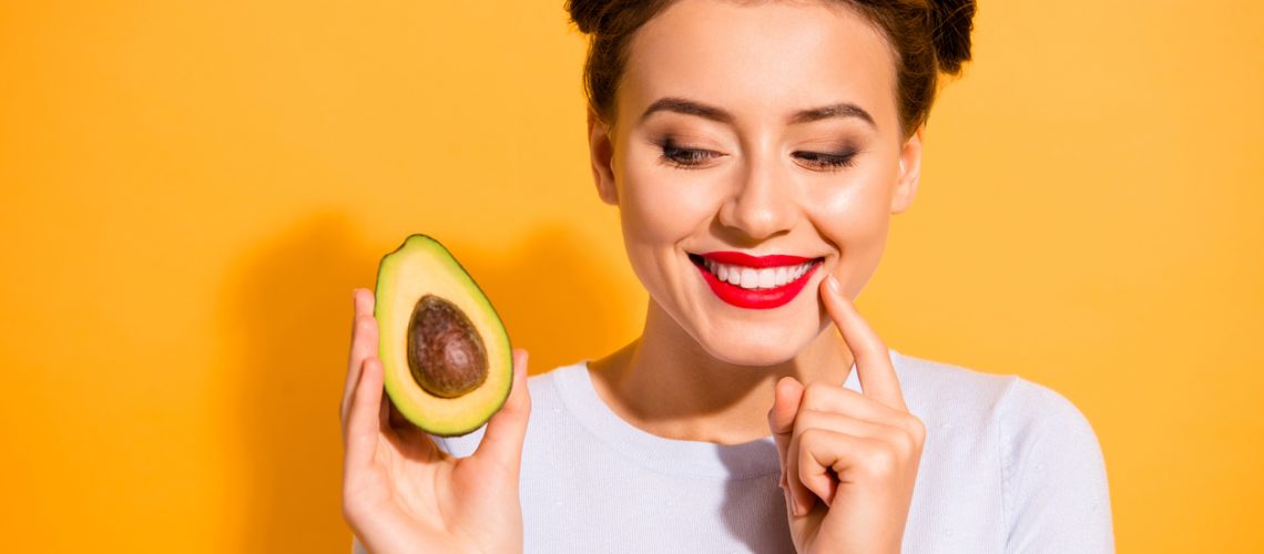 Close up photo beautiful she her lady pretty hairdo hold slice avocado look side with interest hand arm finger chin yummy tasty wear casual white pullover clothes isolated yellow background.