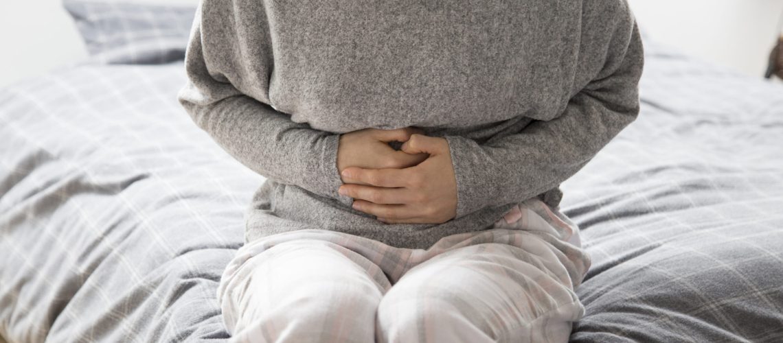 Sick woman in grey homewear sitting on bed, keeping hands on stomach, suffering from intense pain. Illness, stomach ache concept