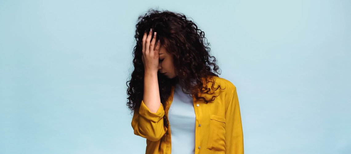 depressed woman with wavy hair covering face with hand isolated on blue