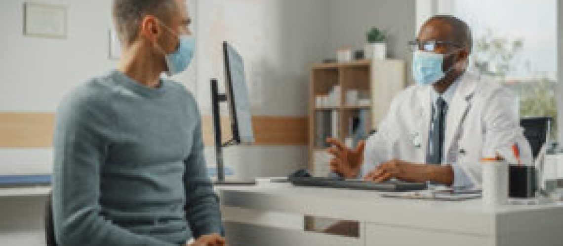 Middle Aged Family Doctor is Talking with Young Male Patient During Consultation in a Health Clinic. Both Wear Face Masks. Physician in Lab Coat Sitting Behind a Computer Desk in Hospital Office.